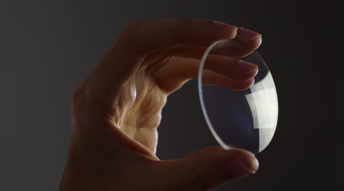 Hand holds an eyewear glass lens, glass prescription lens on dark background
